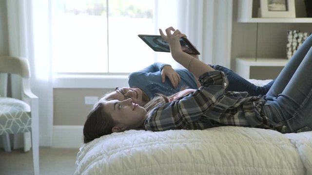 Dolly shot of playful sisters taking selfie through tablet computer while lying in bed at home