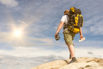 male tourist climbing to the top of the mountain. a large yellow backpack, khaki shorts without a face, binoculars and a map in hand. sky background.