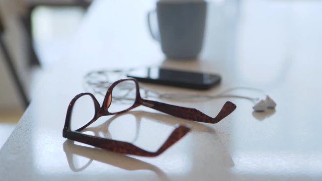 Handheld shot of eyeglasses with smart phone and headphones by coffee cup on table at home