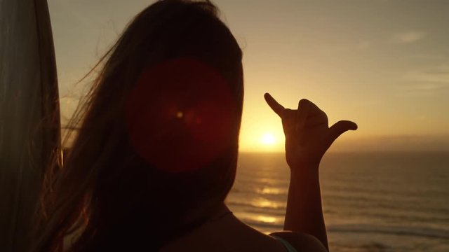 SLOW MOTION CLOSE UP LENSE FLARE: Young surfer girl giving the shaka sign. Peaceful woman enjoying the beautiful nature with wind in her hair making the hang loose hand gesture. The Hawaiian greeting.