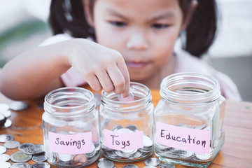 Cute asian little child girl putting coin into glass bottle in the garden. Kid saving money for the...