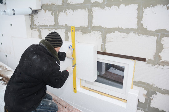 Building contractor insulating House Wall with styrofoam insulation sheets in problem area around window. Worker measuring styrofoam board with spirit level and cutting with knife.