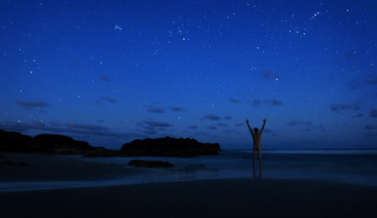 Fototapeta na wymiar Naked man stands alone on beach at night to celebrate the clear sky of stars with hands in the air.