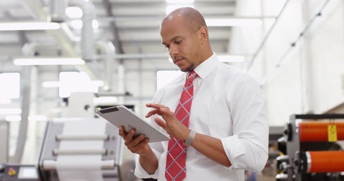 4K Business manager in print factory working on computer tablet, machinery & staff members in background. Slow motion.