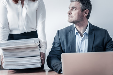 Additional work. Busy working man typing on a laptop and focusing his attention a female coworker standing next to him and holding a pile of business documents.