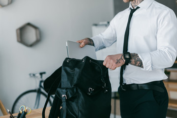 cropped shot of young tattooed businessman putting laptop into backpack