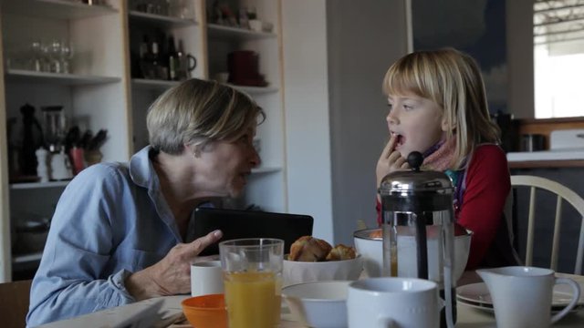 Handheld shot of grandmother with tablet computer talking to granddaughter at breakfast table