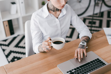 cropped shot of young businesswoman with tattoos holding cup of coffee and using laptop