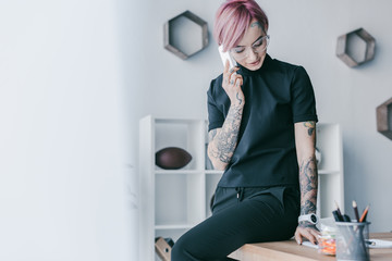 smiling young businesswoman with tattoos talking by smartphone at workplace