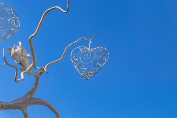 stylized filigree heart hanging on the tree of forged metal with flower on blue sky background
