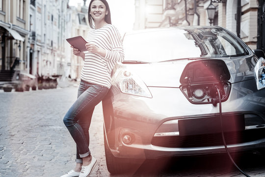 Conserving energy future. Positive minded young brunette smiling cheerfully while leaning on her car and browsing the Internet on her digital tablet during an electric automobile charging session.