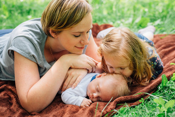 Young mom with her 4 years old daughter and 4 months old baby boy are relaxing and playing on blanket in the summer park