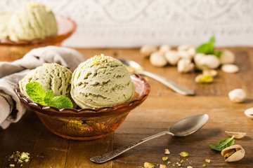 Fototapeta premium Pistachio ice cream scoops with grated pistachos in red glass bowl on old wooden table background. Homemade summer food concept with copy space.