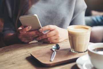 Closeup image of a woman holding and using smart phone with coffee cups on wooden table in cafe