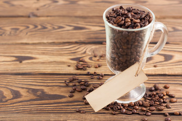 Lot of fried coffee beans in transparent glass for mulled wine with rectangle blank paper on rope near scattered coffee beans on old wooden rustic brown planks