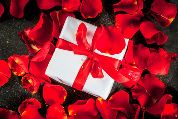 Valentine's day concept, with rose flower petals and white wrapped gift box with red ribbon, on dark stone background, copy space top view