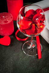 Valentine's day concept, white wrapped gift box with red ribbon, rose flower petals in wine glass, with red candle, on dark stone background, copy space
