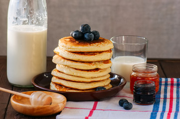 Stack of homemade pancakes with blueberries in a plate, milk and jam preserves in a glass jar.  Rustic style.