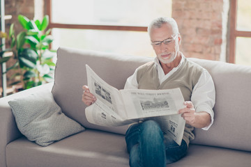 Calm careless atmosphere. Portrait of concentrated confident interested peaceful serious old granddad with white hair sitting on beige sofa with crossed legs reading favorite newspaper