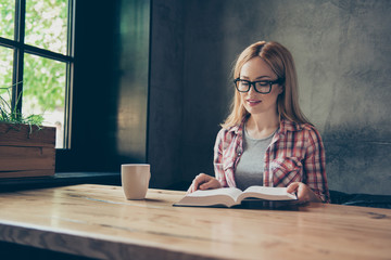 The best source of getting knowledge is reading a book! Smart clever beautiful cute woman is reading a thick book and drinking fresh tea, sitting at the table in a cafe