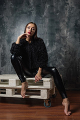 Brunette woman sitting on wooden pallet couch. Sexy female model in elegant black pants and short eco faux fur coat thinking and dreaming