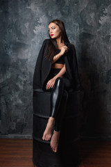 Brunette woman sitting on a barrel. Sexy female sad model in elegant black leather pants and jacket thinking and dreaming