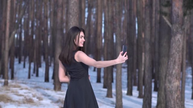 Portrait attractive smiling young brunette girl in a black dress makes selfie on a snowy forest landscape background