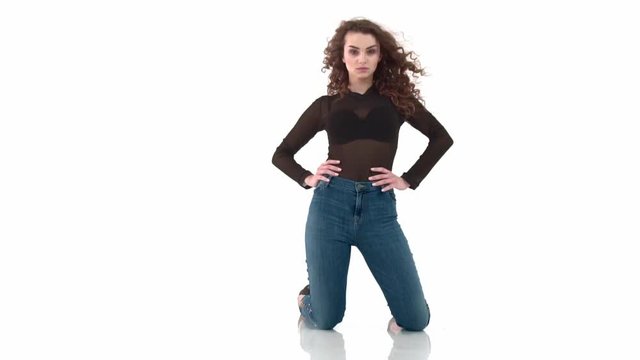 Curly-haired girl in black body and jeans kneeling on a white background slow motion