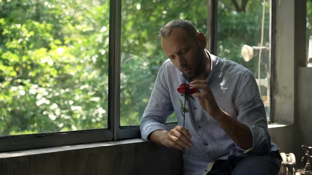Pensive, heartbroken man holding red rose sitting by window at home
