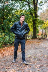 Fototapeta na wymiar Young American man traveling at Central Park, New York in autumn day. Man wearing black leather jacket, jeans, gray casual shoes, hands in pockets, walking on road with trees, fallen leaves on ground