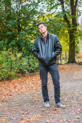 Fototapeta na wymiar Young American man traveling at Central Park, New York in autumn day. Man wearing black leather jacket, jeans, gray casual shoes, hands in pockets, walking on road with trees, fallen leaves on ground,