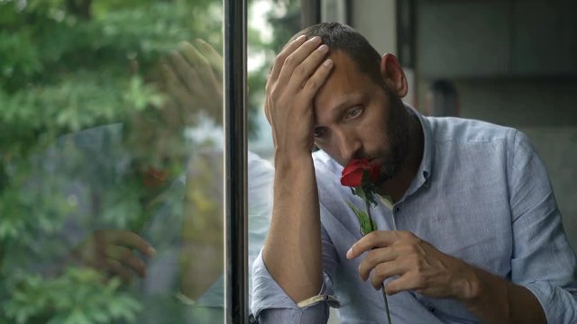 Sad, unhappy man holding red rose sitting by window
