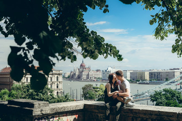 Fototapeta premium Romantic sunny portrait of the sensual couple in love tenderly rubbing noses while sitting on the balcony at the panorama view of Budapest, Hungary.