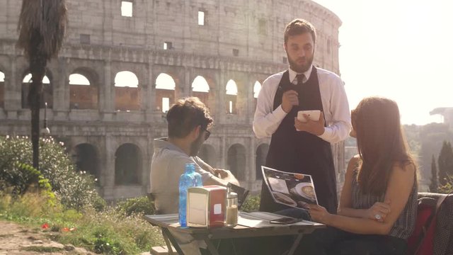 Happy young couple tourists ordering food an drinks to elegant waiter sitting at bar restaurant in front of colosseum in rome at sunset