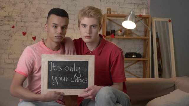 A sad international gay couple is sitting on the couch and holding a sign. It's only your choice. Look at the camera. Home comfort on the background. 60 fps
