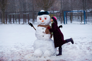 A little cheerful girl kiss funny snowman face. A cute little girl has fun in winter park, wintertime
