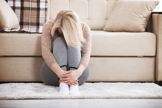 Middle aged barefoot woman sitting at the floor embracing her knees, near sofa at home, her head down, bored, troubled with domestic violence.