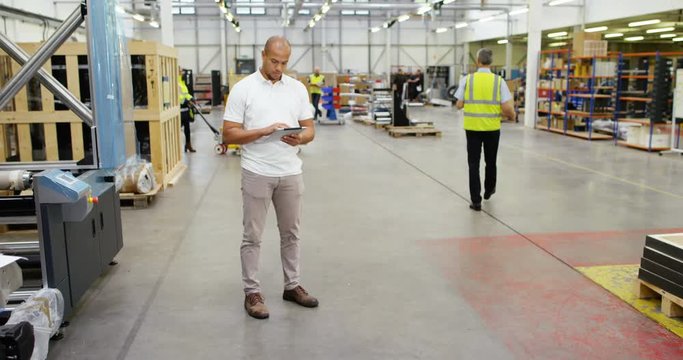 4K Young business manager in factory warehouse looking at tablet computer, with workers busy in the background
