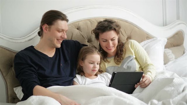 Parents and Their Daughter are Using Tablet Lying on the Bed. Young Pretty Mother with Curly Hair is Laughing.