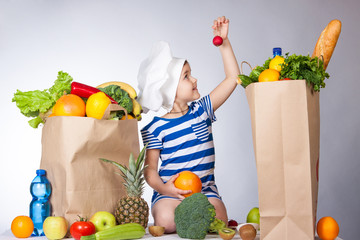 Little happy girl in the hat of the chef with big bags of food. A variety of fresh fruits and vegetables in bags on the table. Health food.