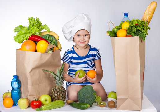 Little happy girl in the hat of the chef with big bags of food. A variety of fresh fruits and vegetables in bags on the table. Health food.