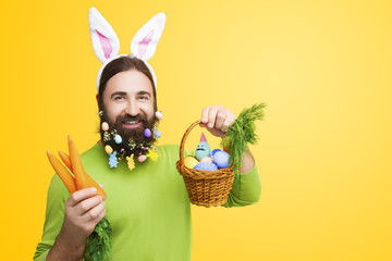 Happy Easter concept. Nice kind muscle man male with beard, white ears of rabbit, carrots and basket with colorful easter eggs and green smiling in green t-shirt isolated on yellow background