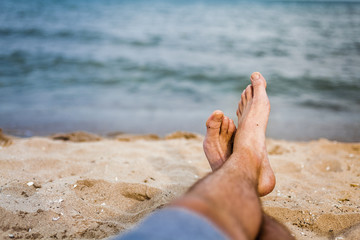 An adult crosses his sandy feet on the beach while the water is faded in the background on a summer afternoon.