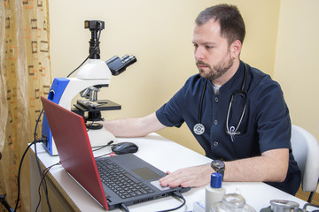 Young handsome doctor checking the blood sample on his microscope in his medical office