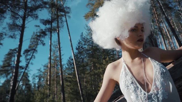 Woman like a fallen angel without wings is standing in a forest and looking in a camera. She has a white curly hair and white lace dress, close-up of face