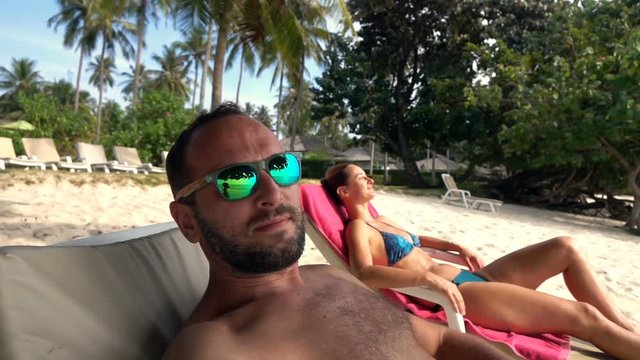 Happy man taking selfie, video on sunbed on beach, super slow motion
