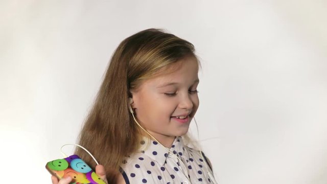 Child with earflaps dancing at studio background. The girl listens to music on the smartphone. The kid has loose long hair. Shooted on a gray white background.