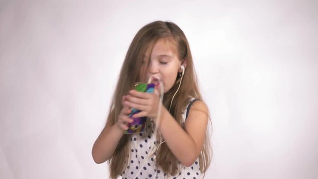 Child with earflaps dancing at studio background. The girl listens to music on the smartphone. The kid has loose long hair. Shooted on a gray white background.