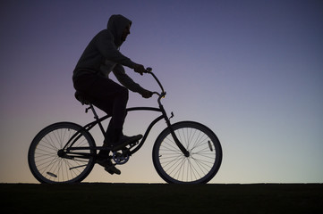 Fototapeta na wymiar Silhouette of a man in a hoodie riding a cruiser bike in front of a purple sunset sky