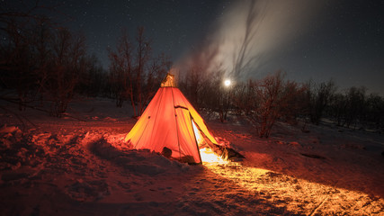 A night photo of arctic landscape with a tee-pee like tent illuminated by a fire inside projecting light out on the snow, Starry clear sky with half moon in the background. (Long exposure time)